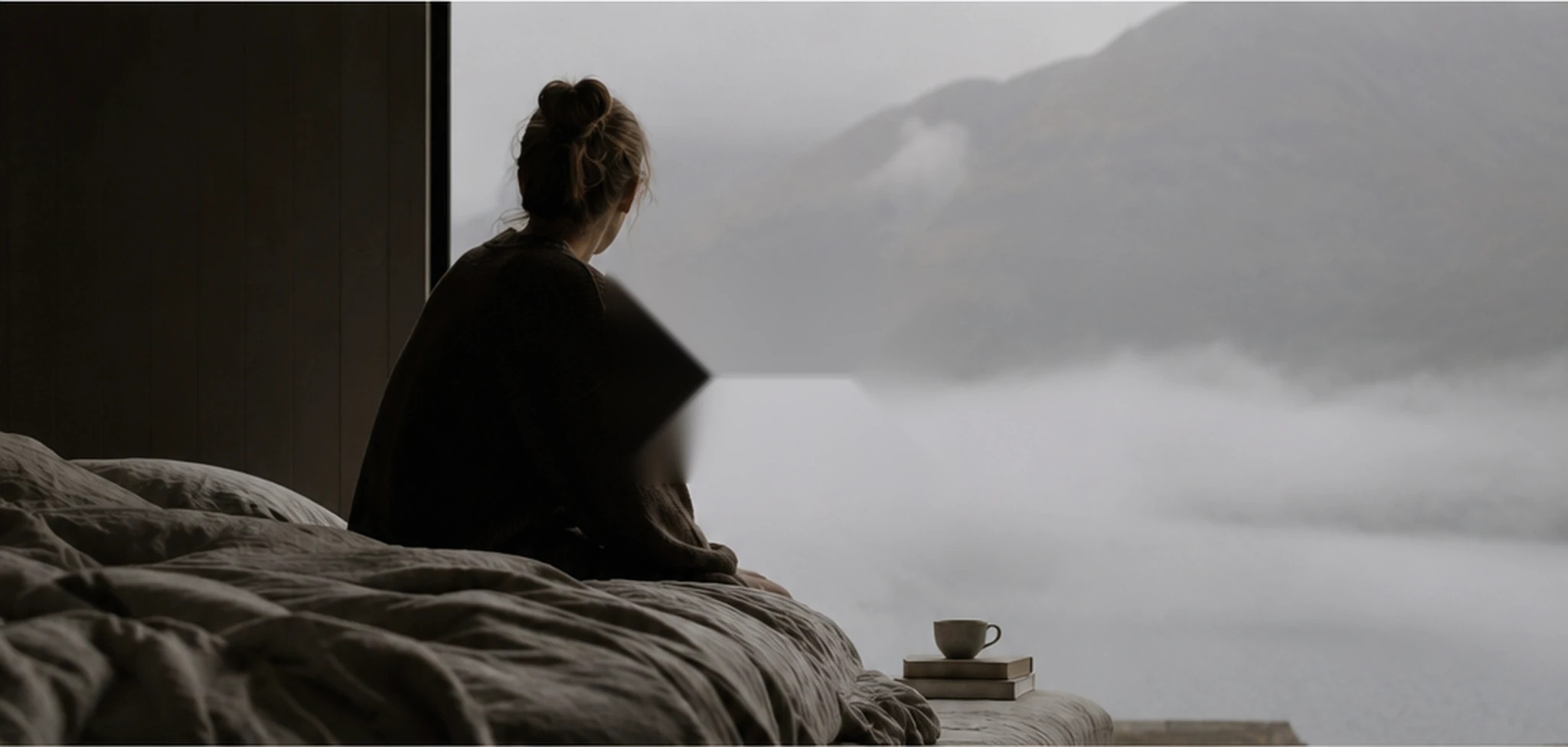 A woman sitting quietly on an unmade linen bed overlooking mist-covered mountains
